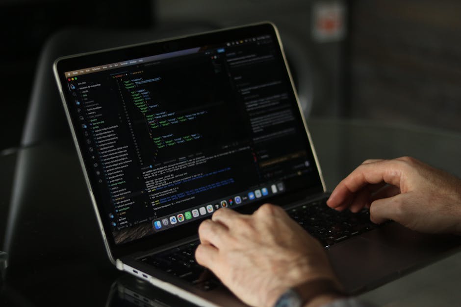 A person using a laptop computer with a dark-themed code editor open on the screen, displaying lines of colorful programming code. The laptop is placed on a dark, reflective surface, and the person's