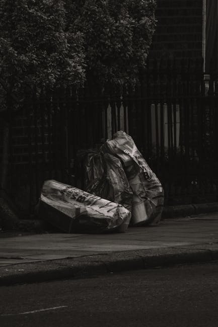 A person disposing of brown paper bags filled with rubbish into a white recycling or waste collection bin, positioned on a wooden surface. The person's arm and hand are visible, holding one paper bag